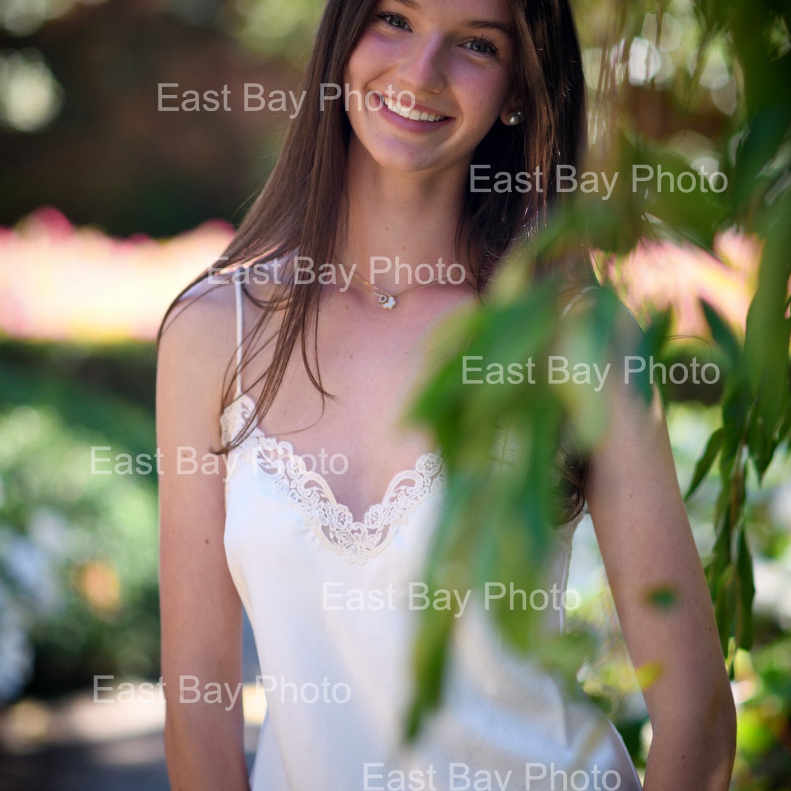 This portrait was taken at Filoli Historic House & Garden, in Woodside, California. It was taken available light only because this location does not allow the use of a flash or other photography accessories. The young woman is wearing a white blouse and dark blue jeans. She tilts her head to the left and smiles while her open hands rest against her tilted hips. In the foreground, soft-focused green leaves, obscure portions of her blouse while the background is filled with mottled light from other plants and flowers.